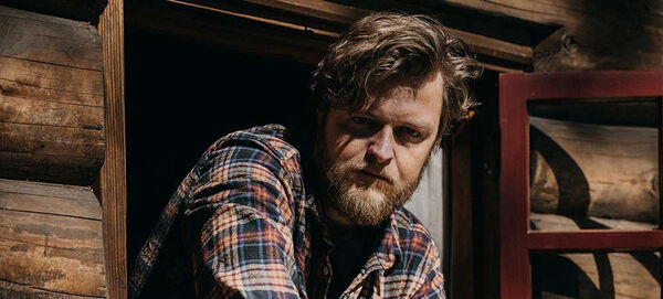 Man met een baard in een geblokt overhemd kijkt vanuit een raam van een houten huis.