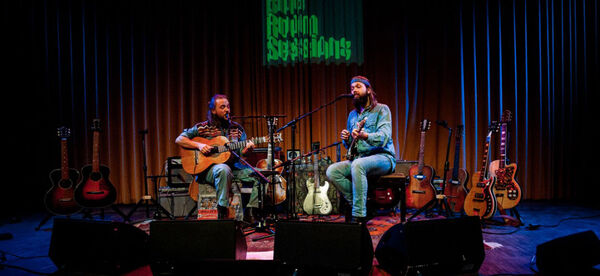 Twee muzikanten met gitaren op een podium, omringd door diverse gitaren, met een sfeervolle achtergrondverlichting.