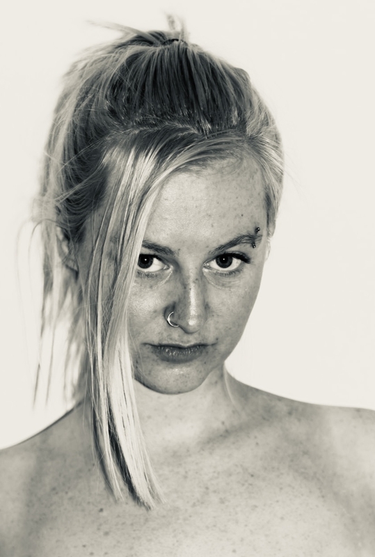 Portret van een jonge vrouw met blond haar, recht in de camera kijkend, met een neutrale achtergrond.