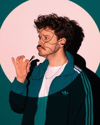 Jongeman in sportoutfit met een stijlvolle krul, poses met een hand aan zijn gezicht tegen een kleurrijke achtergrond.
