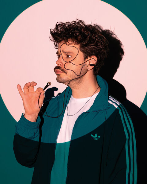 Jongeman in sportoutfit met een stijlvolle krul, poses met een hand aan zijn gezicht tegen een kleurrijke achtergrond.