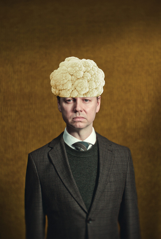 Een man in een donker pak met een bloemkoolsnede als haarstijl, tegen een bruin achtergrond.