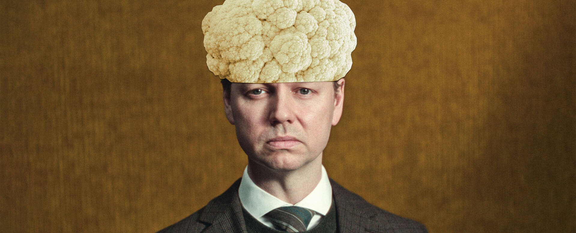 Man in formele kleding met een grote, witte hoed van pluis bovenop zijn hoofd, op een achtergrond in warme kleuren.