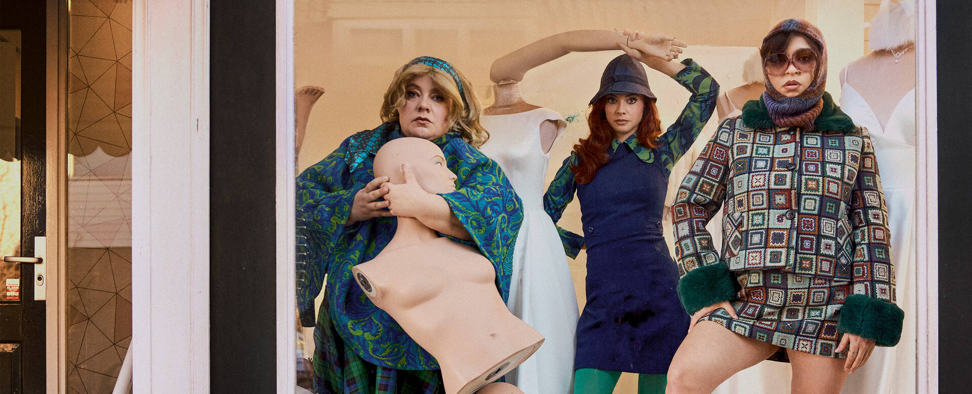 Drie modellen in creatieve outfits poseren voor een moderne achtergrond, met een pop als accessoire.