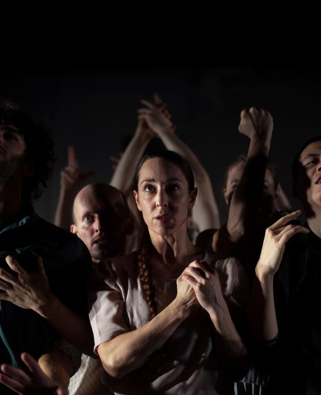 Een groep mensen in een theatrale houding, met gefocuste gezichten en opgestoken handen, in een donkere omgeving.