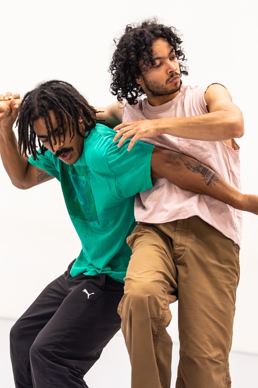 Twee mannen in casual kleding zijn aan het dansen in een lichte ruimte, met expressieve lichaamshoudingen.