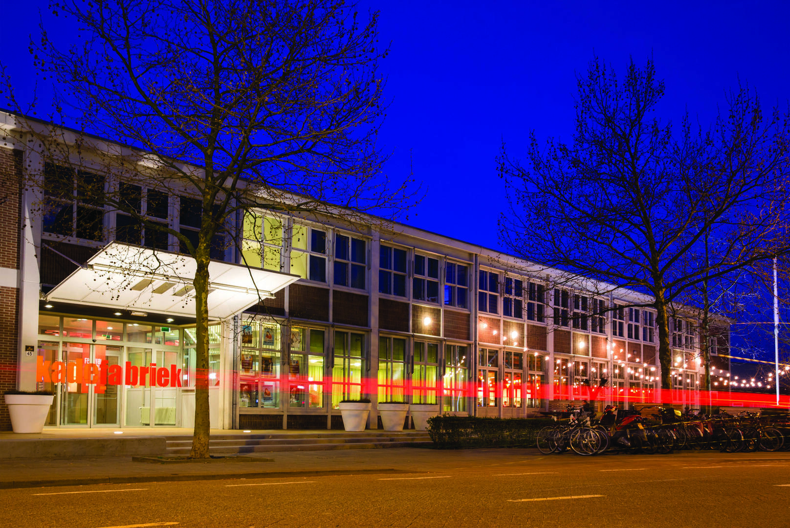 Verkadefabriek in het donker, met verlichting en bomen vooraan.