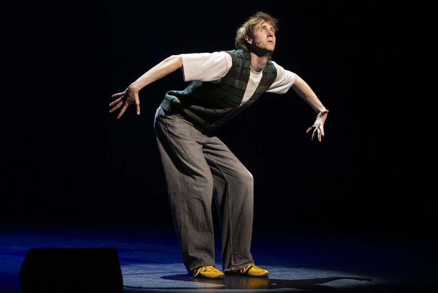 Een performer in een grijs kostuum en gele schoenen, die een expressieve danspose aanneemt op een donker podium.