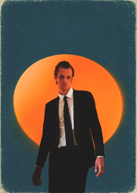 Een man in een zwart pak staat voor een grote, oranje zon op een blauwe achtergrond.