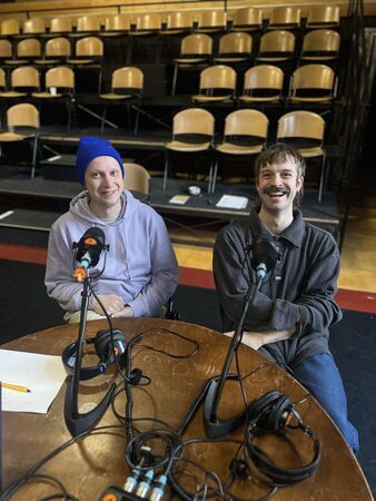 Twee mannen zitten aan een tafel met microfoons in een theateromgeving, beide lachen en dragen hoofdtelefoons.