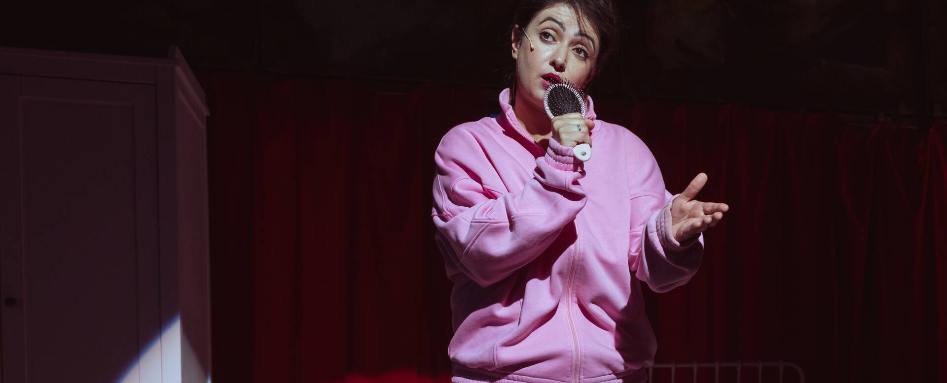 Vrouw in een roze sweater staat op een podium en houdt een microfoon vast terwijl ze spreekt.