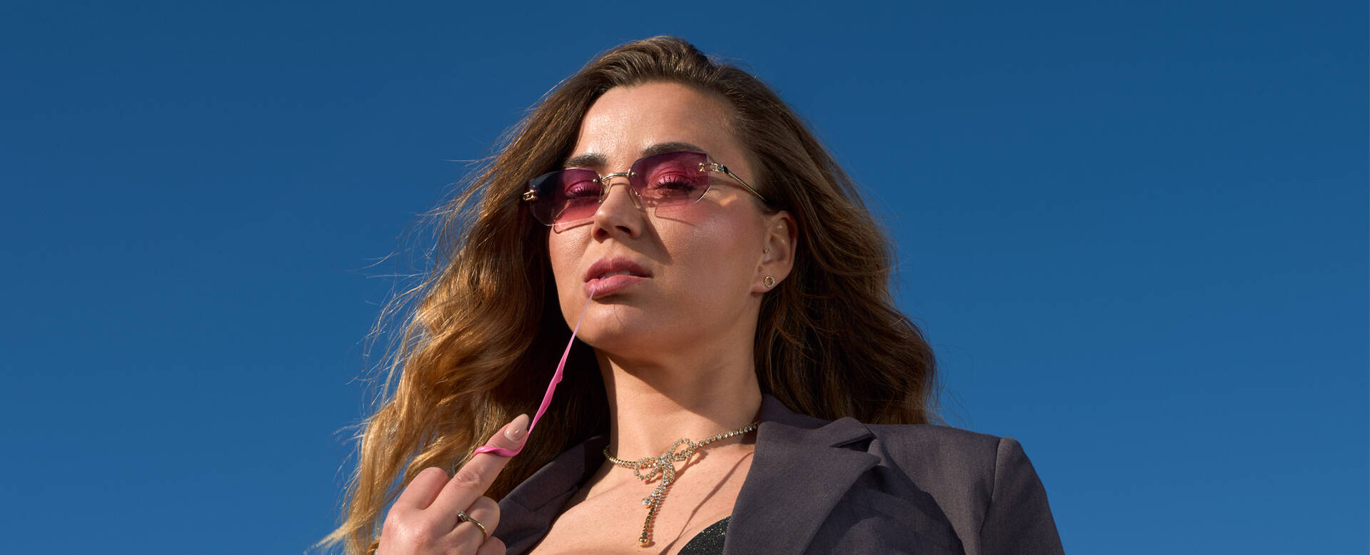 Vrouw met lange, golvende haren in een zonnige omgeving, draagt een donkergrijze blazer en roze zonnebril.