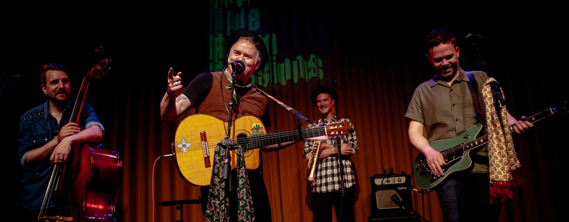 Een bandleden optreden op een podium, met een zanger met een gitaar en muzikanten op contrabas en gitaar.