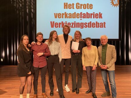 Groep mensen op een podium, poseert voor het 'Verkiezingsdebat' met een achtergrond met tekst.