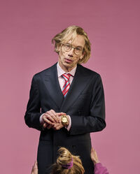 Man in een zwart pak met bril en een roze achtergrond, terwijl een kind zijn handen vastpakt.