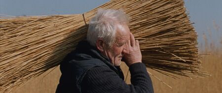 Een oudere man draagt een grote bundel stro op zijn schouder in een open veld.