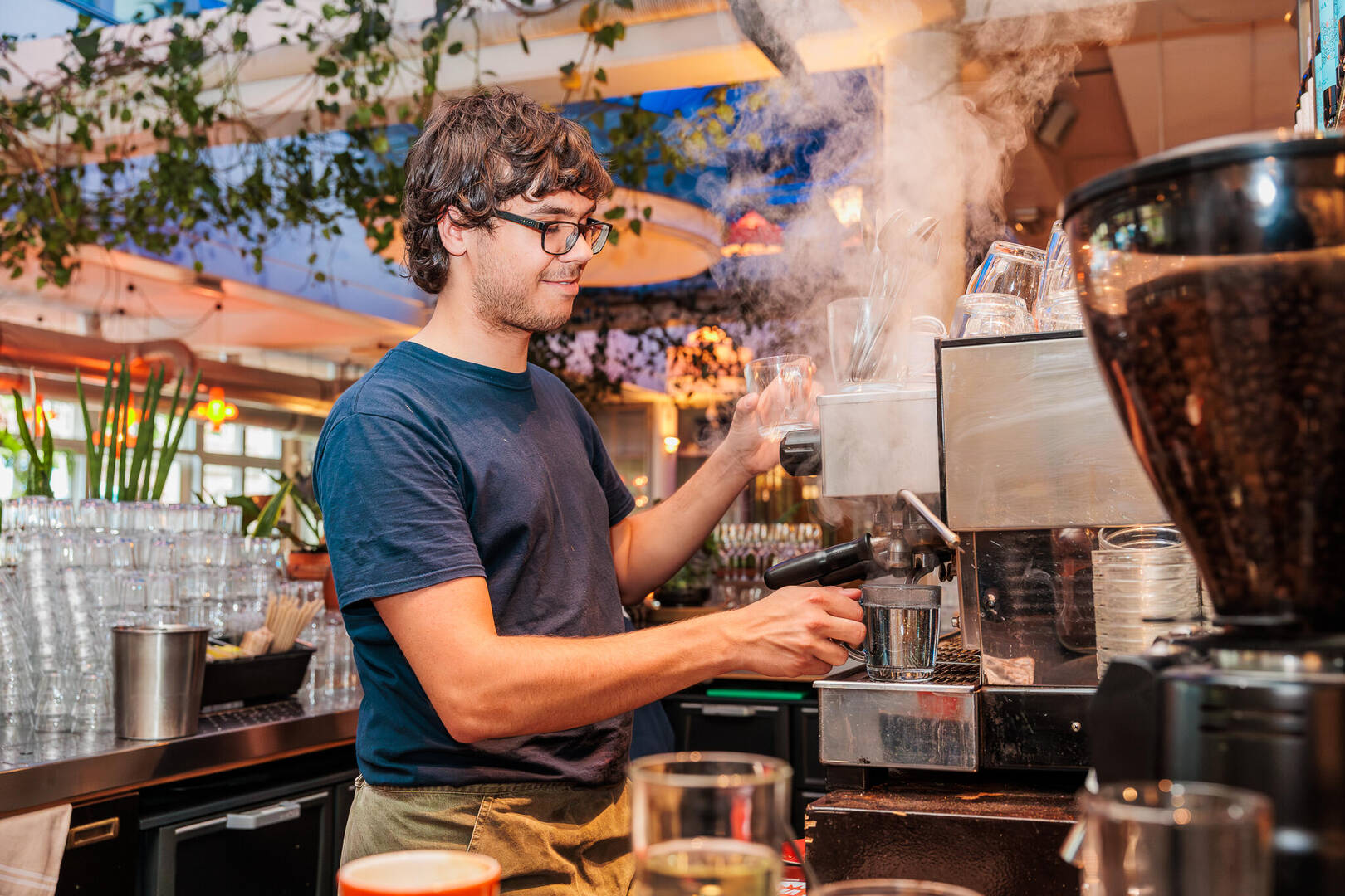 Barista aan het werk met een koffiemachine, omgeven door planten en een levendige atmosfeer.
