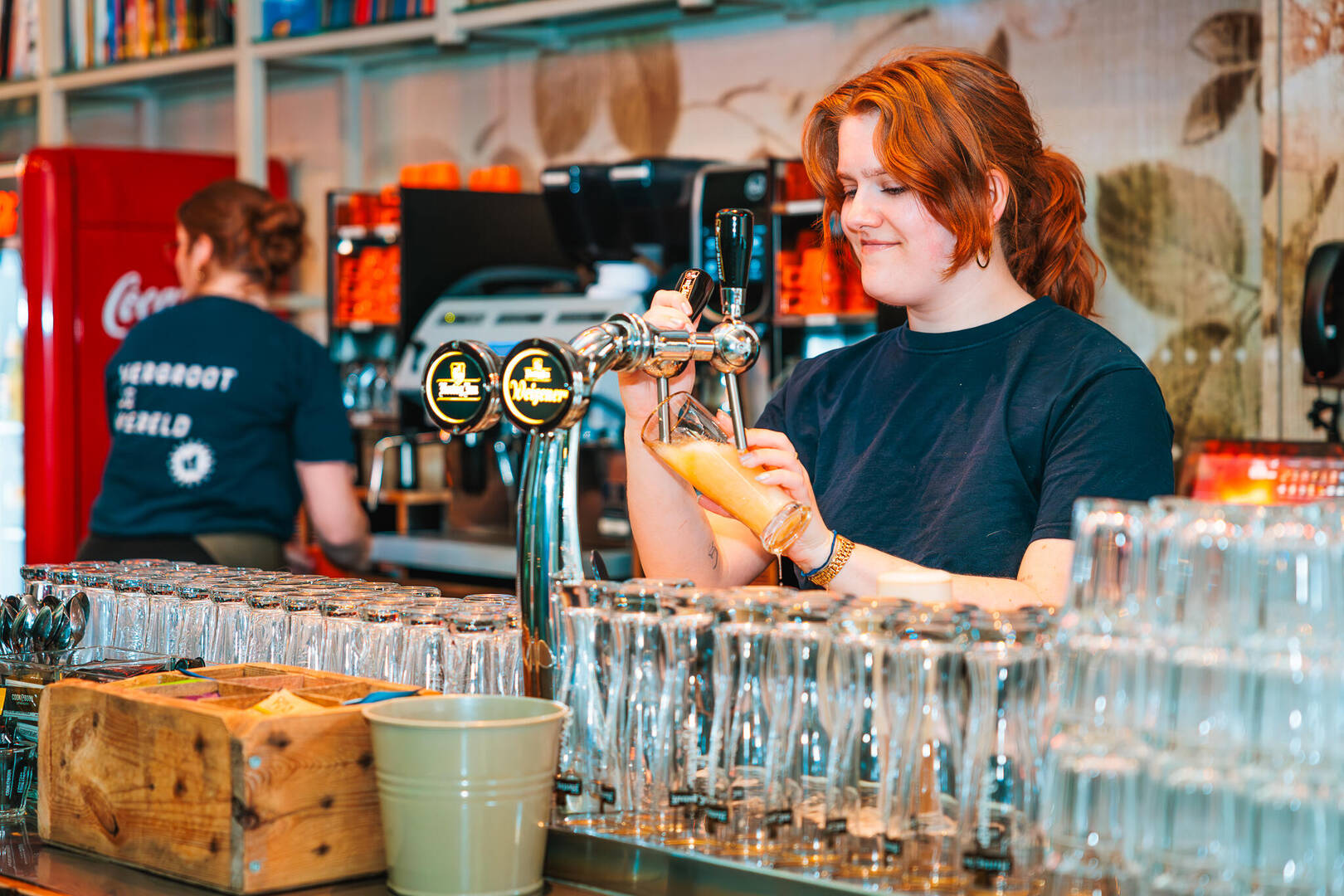 Een serveerster tapt een drankje aan de bar, terwijl een collega achter haar bezig is met het koffieapparaat.