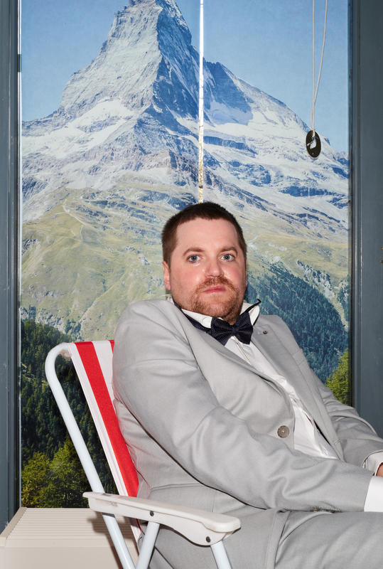 Man in een grijs pak en vlinderdas zit op een stoel voor een bergachtig landschap.