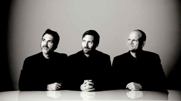 Drie mannen in zwarte kleding zitten aan een tafel, met een neutrale achtergrond en een serious uitstraling.