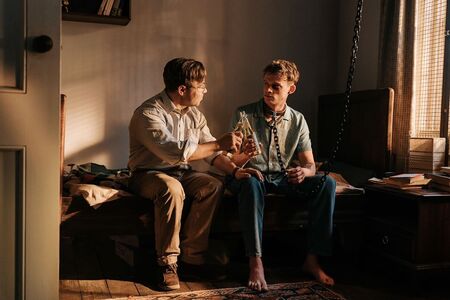 Twee jonge mannen zitten op een bed in een sfeervolle kamer, in gesprek met een fles in de hand.