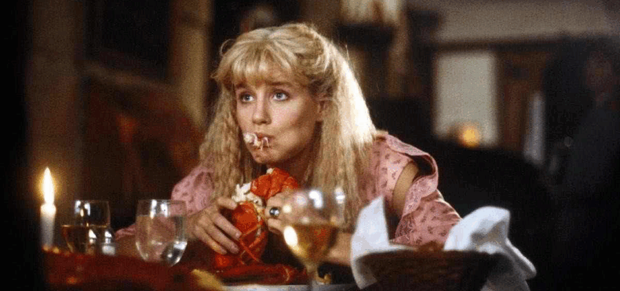 Een vrouw in een restaurant eet een kreeft, met een verraste uitdrukking op haar gezicht en een glas witte wijn naast haar.