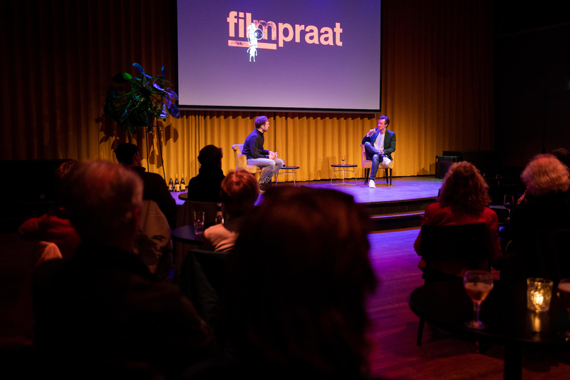 Twee sprekers in gesprek op een podium tijdens een evenement, met het logo van "filmpraat" op de achtergrond.