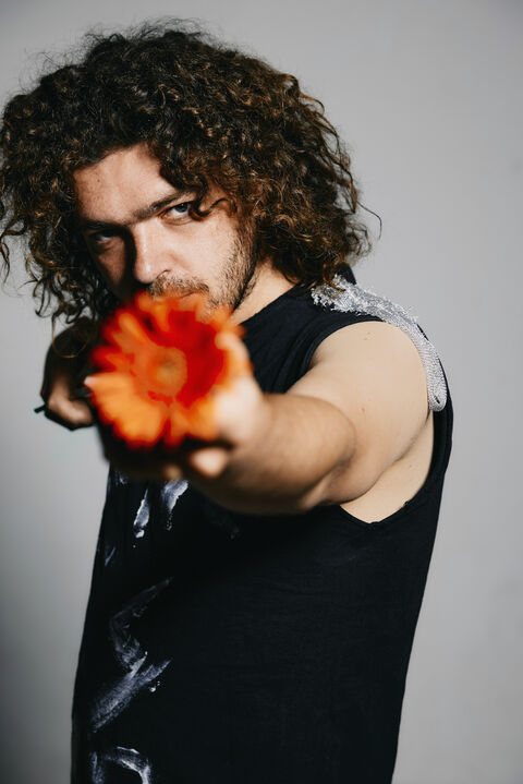 Man met krullend haar in zwart shirt pointed een oranje bloem naar de camera, met een intense blik.