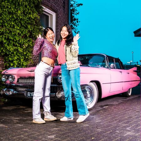 Twee jonge vrouwen poseren vrolijk naast een roze vintage auto op straat.