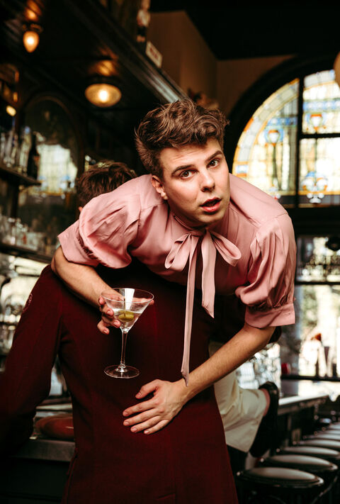 Een persoon in een roze blouse staat op de rug van een andere persoon en houdt een cocktail vast in een café.