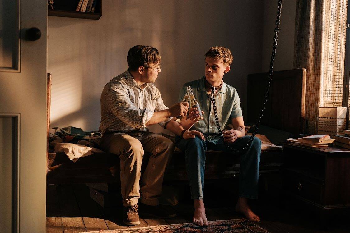 Twee jonge mannen zitten op een bed in een sfeervolle kamer, in gesprek met een fles in de hand.