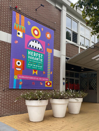 Kleurige poster met de tekst "Herfstvariant" aan de buitenmuur van een gebouw, omgeven door bloempotten.