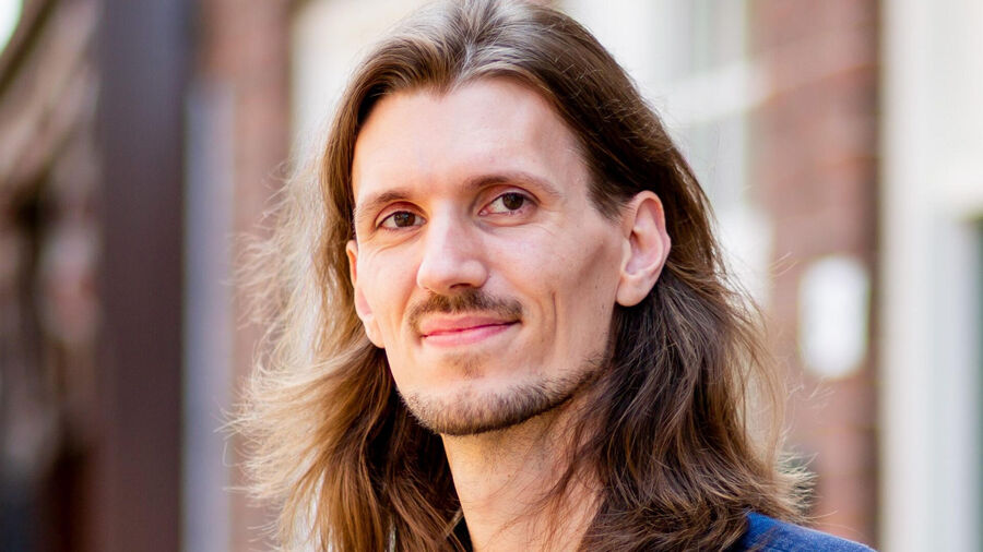 Portret van een man met lang, steil haar en een lichte glimlach, staand voor een achtergrond van bakstenen muren.