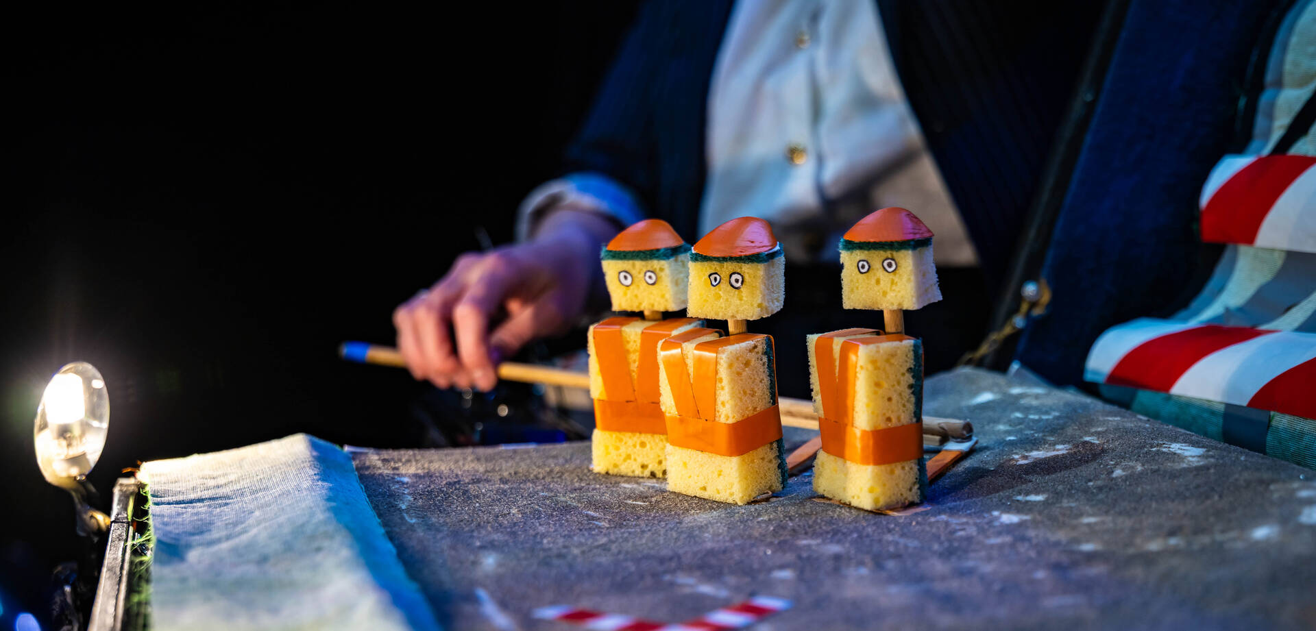 Een hand met een stokje voor houten figuren van huisjes op een podium, bij een theater- of poppenvoorstelling.