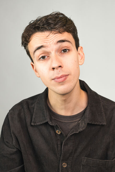 Jongeman met bruin haar en een neutrale blik, gekleed in een donkere trui en shirt, staand voor een lichte achtergrond.