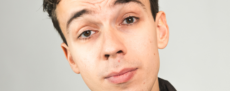 Portret van een jongeman met een neutrale gezichtsuitdrukking en korte, donkere haren, tegen een lichte achtergrond.