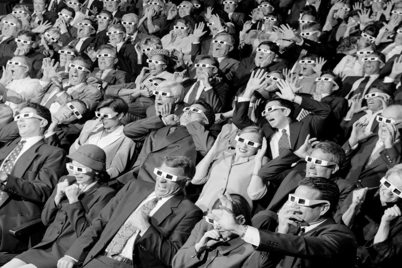 Een zwart-wit afbeelding van een menigte mensen in het theater, allemaal met 3D-brillen op.