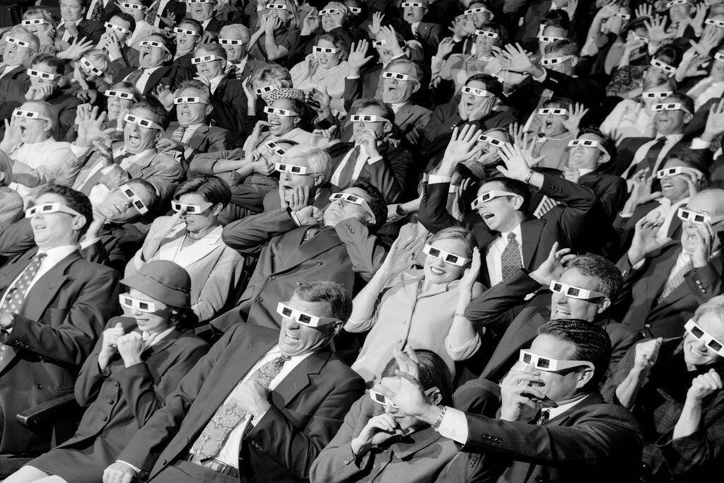 Een zwart-wit afbeelding van een menigte mensen in het theater, allemaal met 3D-brillen op.