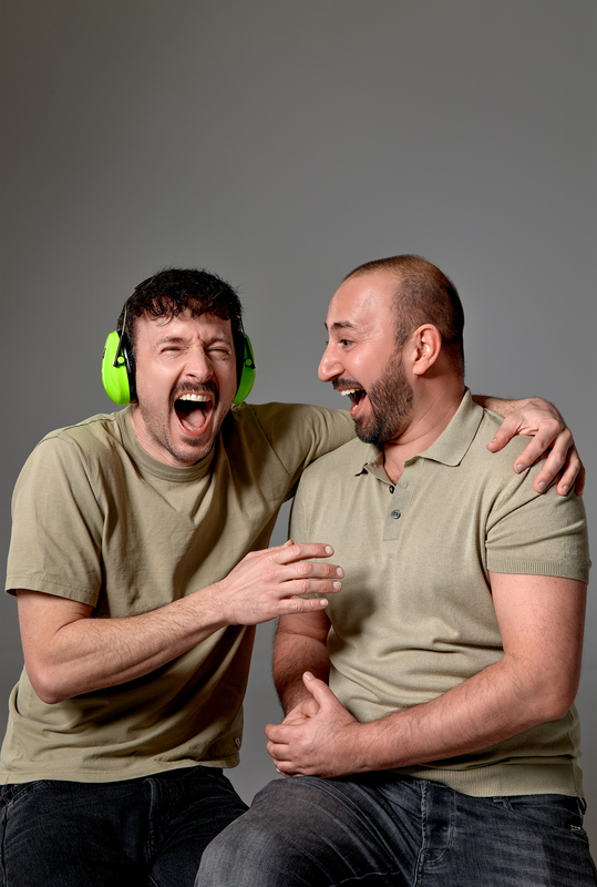 Twee mannen met groene koptelefoons lachen en genieten samen in een neutrale omgeving.
