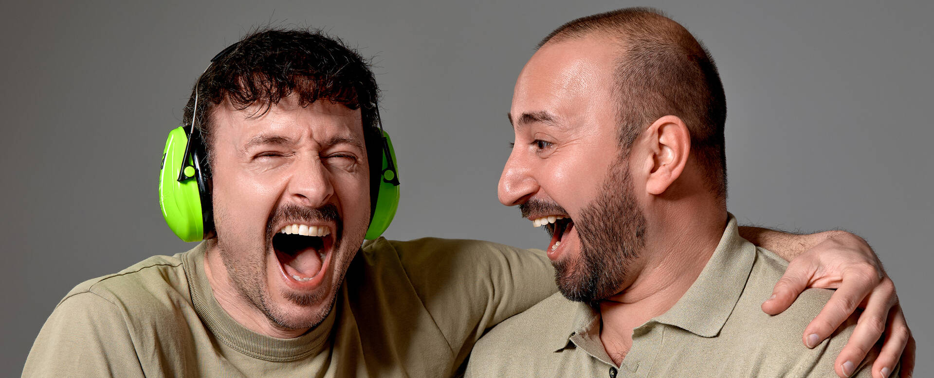 Twee mannen lachen breeduit, met een man die groene koptelefoon draagt, tegen een neutrale achtergrond.