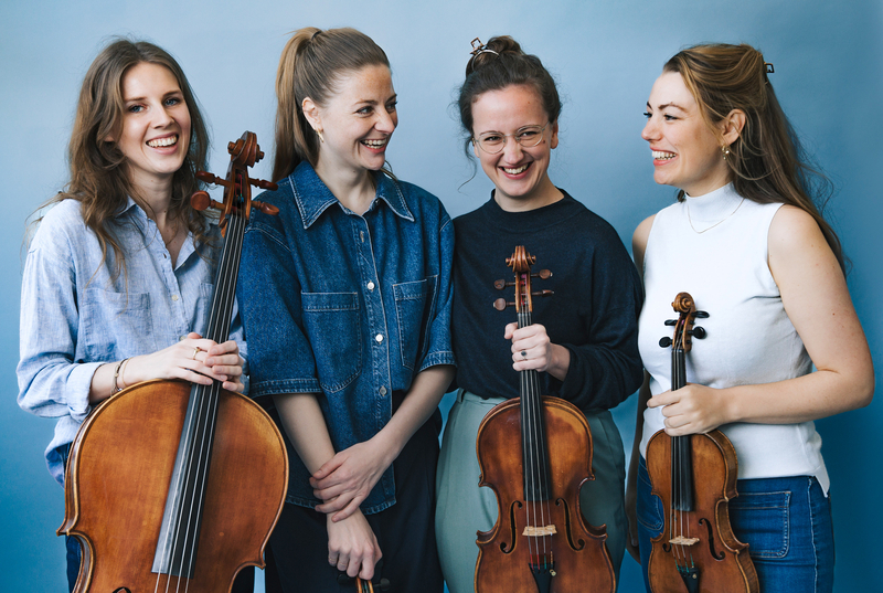 Vier vrouwen staan samen, elk met een strijkinstrument, lachend voor een blauwe achtergrond.