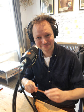 Man met headphones aan de microfoon, lachend en bezig met een potlood of pen in een creatieve omgeving.