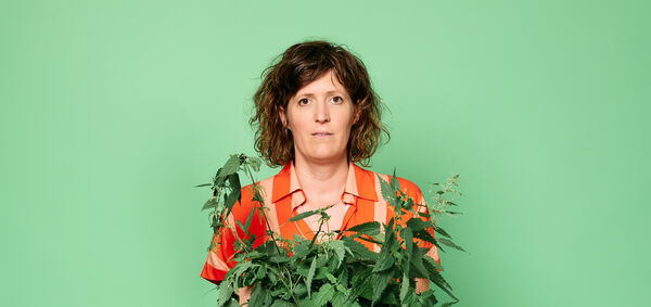  vrouw met krullend haar in een kleurige blouse houdt een plantenpot voor zich tegen een groene achtergrond.