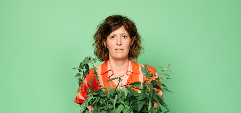  vrouw met krullend haar in een kleurige blouse houdt een plantenpot voor zich tegen een groene achtergrond.