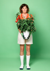 Persoon met krullend haar in een kleurrijke outfit houdt een grote plant vast voor een groene achtergrond.