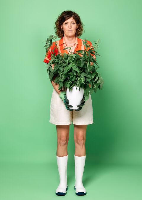 Persoon met krullend haar in een kleurrijke outfit houdt een grote plant vast voor een groene achtergrond.