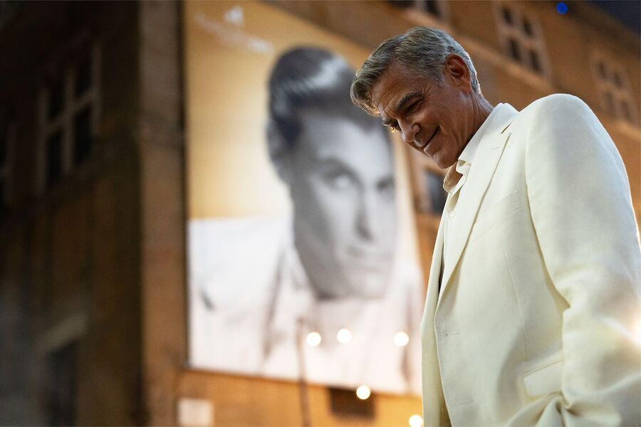 Man in witte blazer staat onder een grote poster van een klassieke filmacteur op een stadsachtergrond.