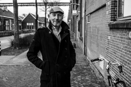 Man in een zwarte jas en pet staat op straat naast een fiets, met gebouwen op de achtergrond in zwart-wit.