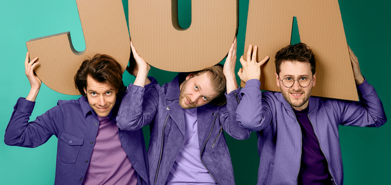 Drie mannen in paarse shirts houden grote kartonnen letters omhoog, die samen het woord "JA" vormen, tegen een groene achtergrond.