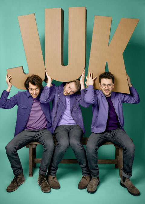 Drie mannen zitten op stoelen en houden een groot kartonnen bord met het woord "JUK" boven hun hoofd.